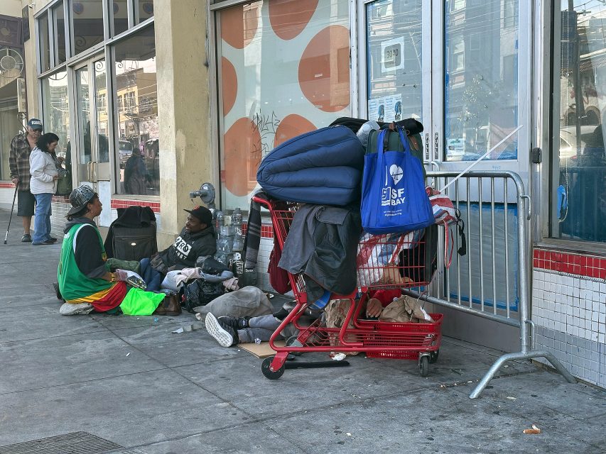 Two people sit on a sidewalk next to a shopping cart filled with belongings outside a building with large windows and a blue SF Bay bag.