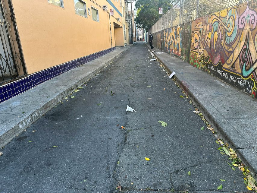 A narrow, empty alleyway with litter scattered on the ground, graffiti on the right wall, and tan buildings on the left side.