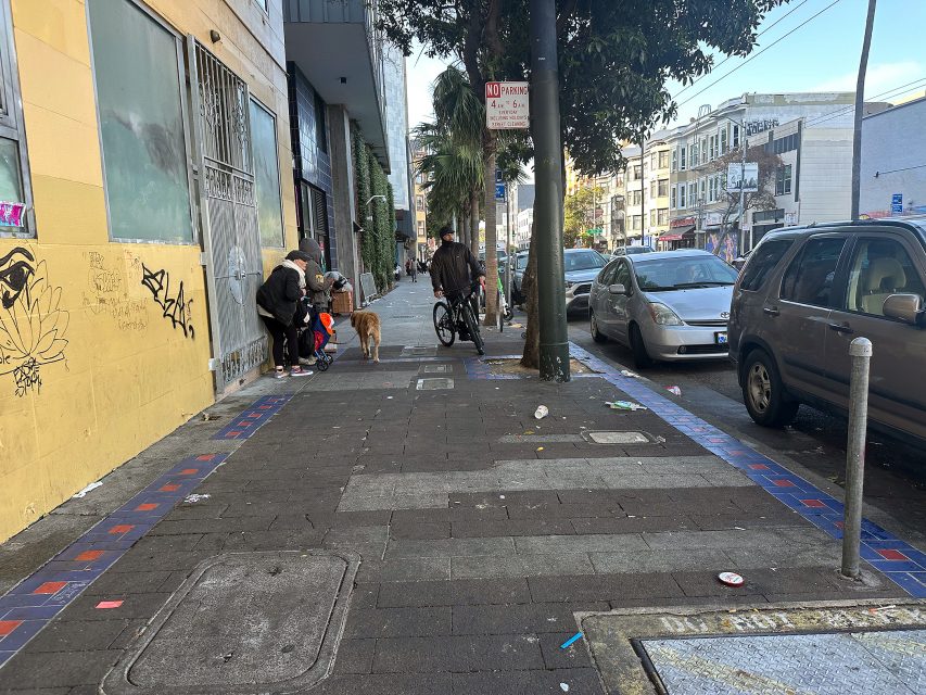 A city sidewalk with scattered litter, parked cars, several people standing near a building, and a person walking a dog.