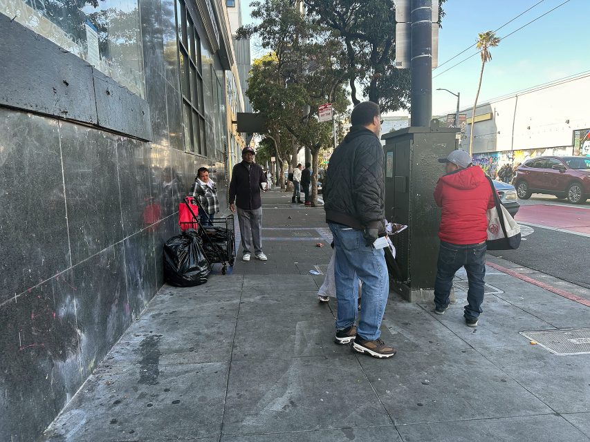 Several people stand and walk on a city sidewalk next to a building, with bags and utility boxes nearby; trees and parked cars line the street.