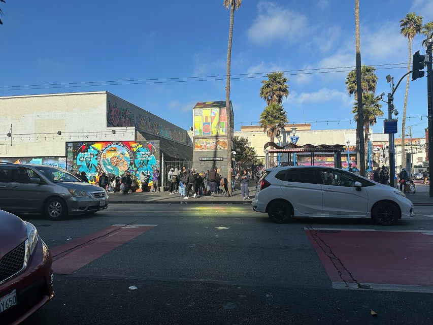 A group of people gathers on a city sidewalk near colorful murals and palm trees, with cars passing on the street in front.