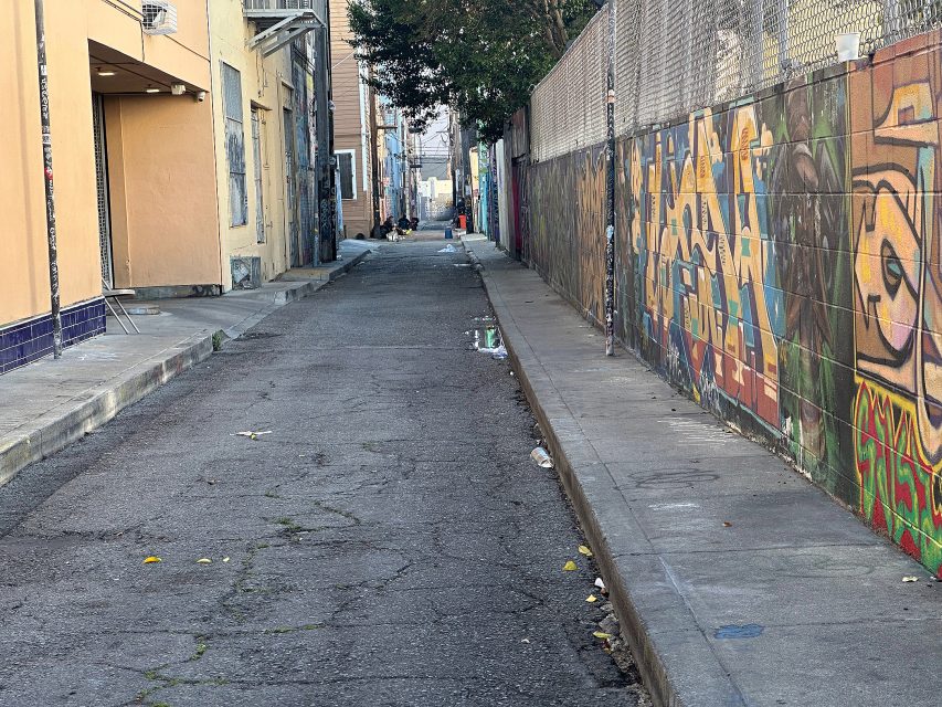A narrow urban alleyway with graffiti on the right wall, litter scattered on the ground, and buildings lining both sides under daylight.
