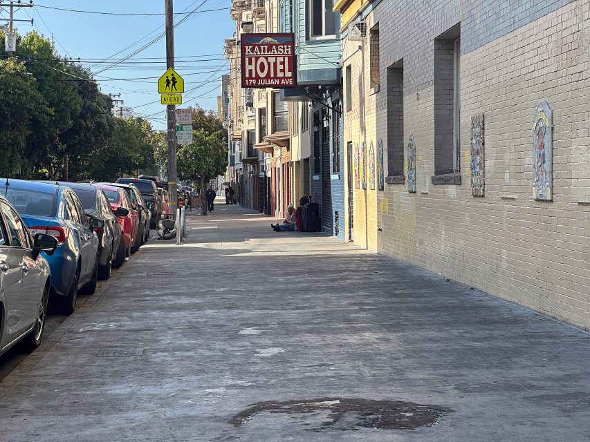A city sidewalk with parked cars on one side and a row of buildings, including the Kailash Hotel, on the other; a pedestrian is visible in the distance.