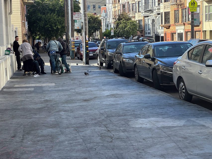 A group of people gather near a building on a city sidewalk next to parked cars; residential buildings line the street.