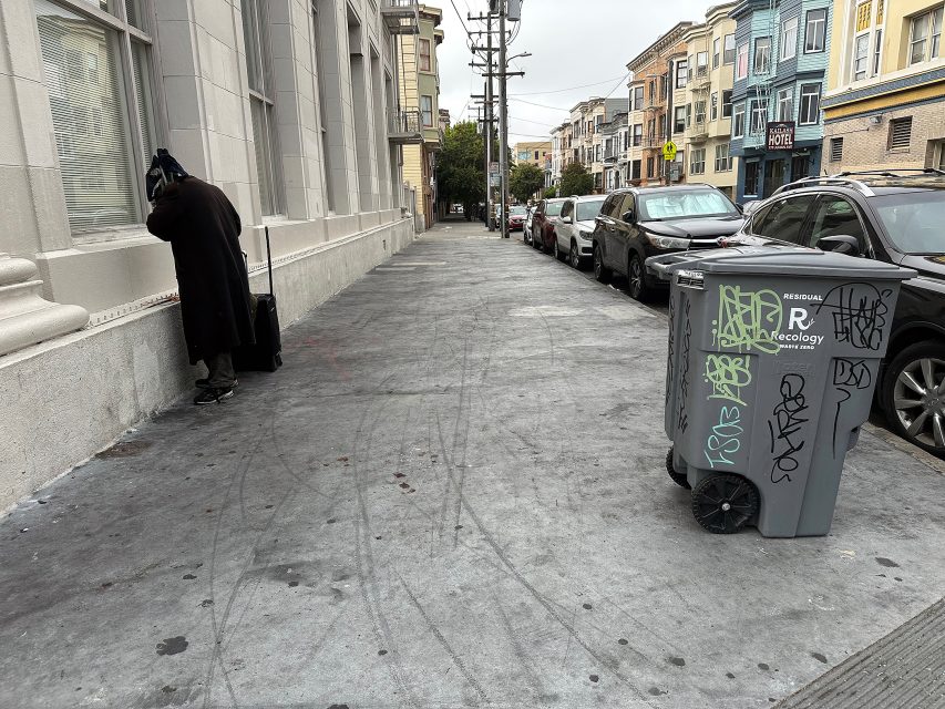 A person stands near a building on a city sidewalk with a suitcase; cars are parked along the street and a graffiti-covered trash bin is in the foreground.
