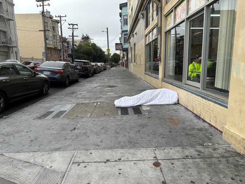 A white blanket or comforter lies unattended on a city sidewalk next to a building, with parked cars and a person visible through a window.
