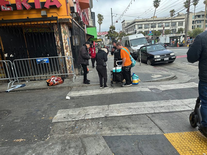 A group of people stand around a table with supplies on a city sidewalk near a corner store and parked cars; buildings and palm trees are visible in the background.