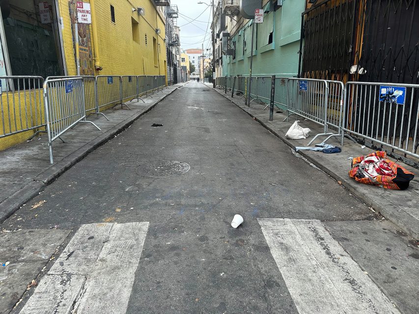 A narrow urban alley lined with metal barricades, scattered litter, and bags on the right side, with yellow and green buildings on either side.