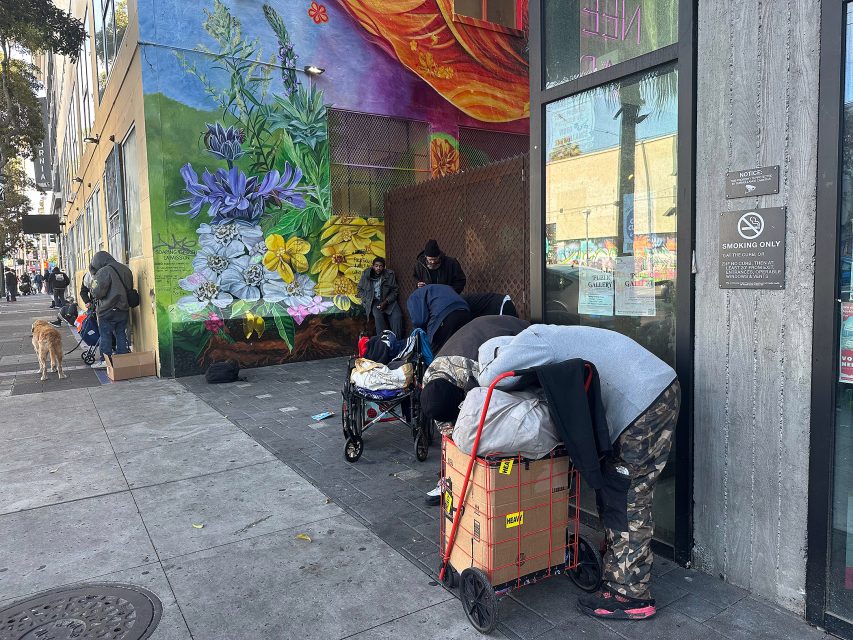 People with carts and belongings gather on a city sidewalk near a colorful mural and a building entrance with a "No Smoking" sign.