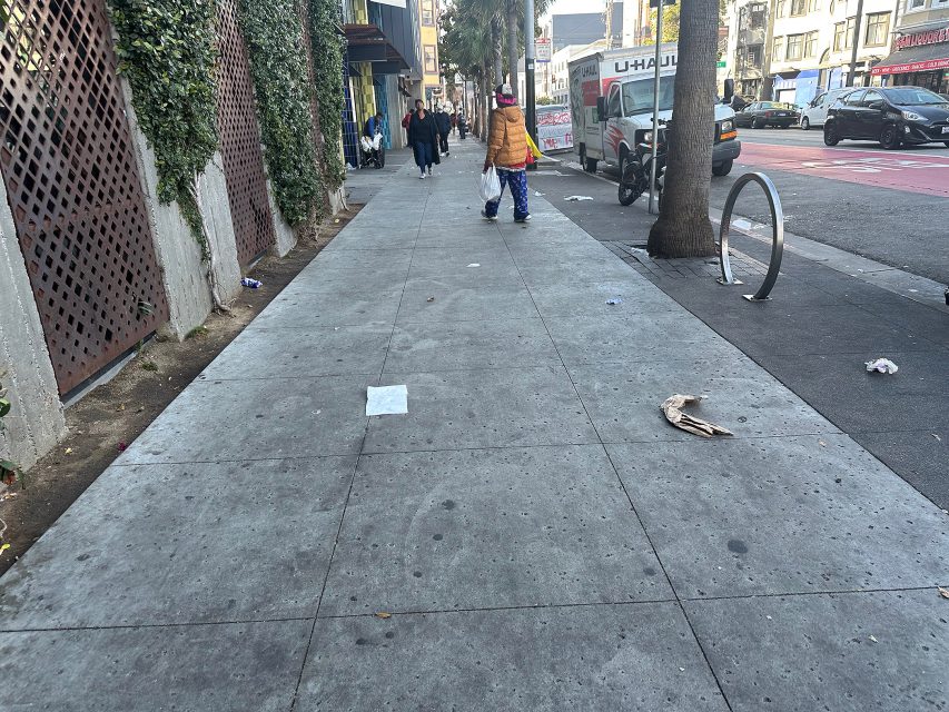 A person carrying bags walks on a city sidewalk scattered with litter, with buildings, parked cars, and pedestrians in the background.