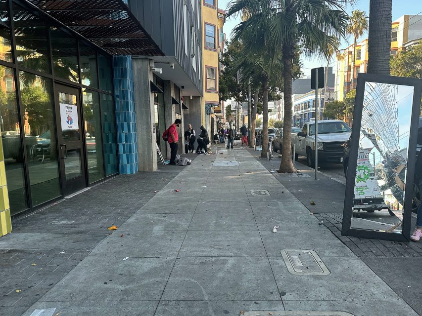 People stand and walk along a city sidewalk with scattered litter, next to a building with glass doors and a large broken mirror on the ground. Cars are parked along the street.