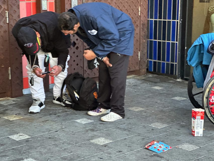 Two people stand on a city sidewalk, focused on something in their hands. A backpack, a bicycle, and litter are visible nearby.