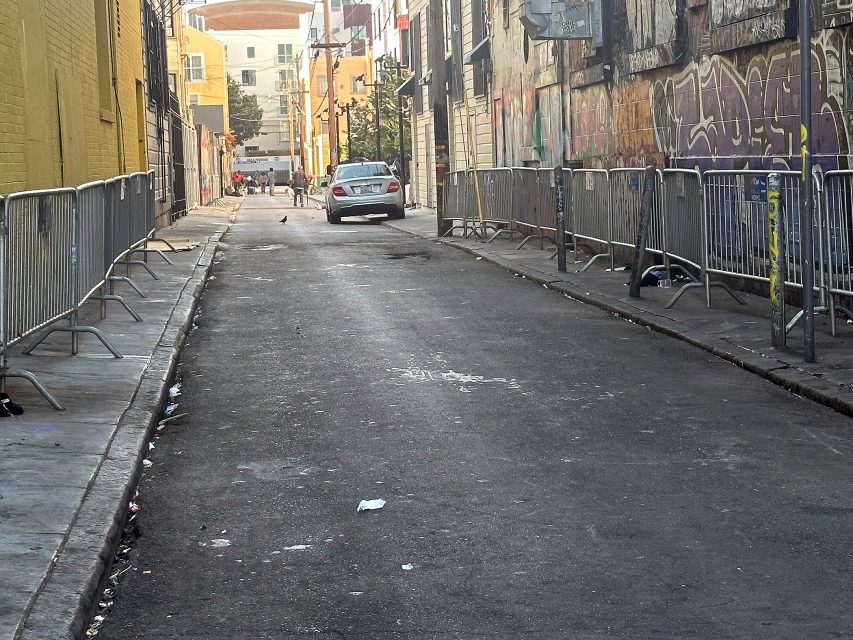 A narrow city alley with graffiti-covered walls, metal barricades along the sides, some litter on the ground, and a parked car near the end of the alley.