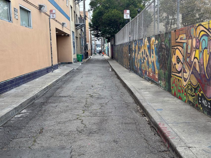 Narrow urban alleyway with cracked pavement, graffiti-covered wall on the right, beige building on the left, and no parking signs visible.