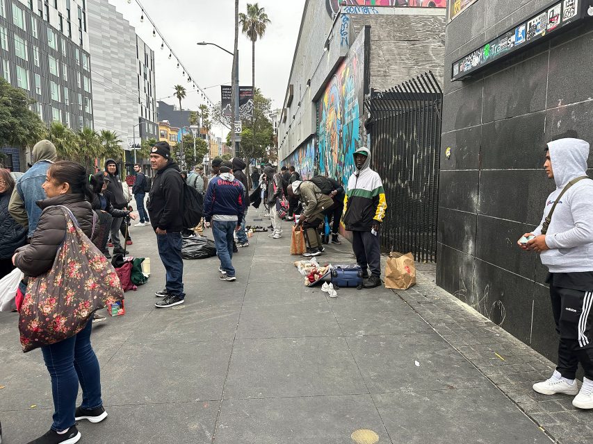 A group of people stand and gather along a city sidewalk lined with graffiti walls, bags, and various belongings on the ground.