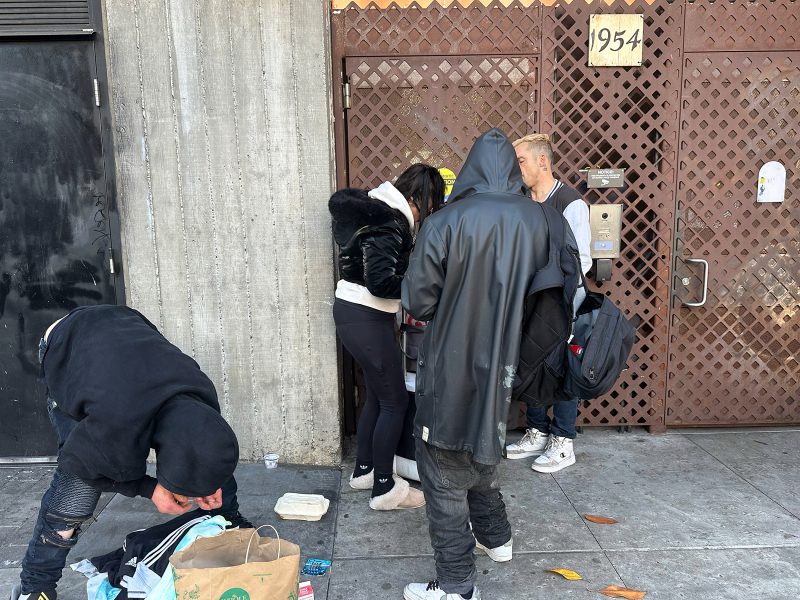Five people stand or crouch on a sidewalk near a gated door marked 1954; some are interacting, with bags and personal items on the ground nearby.