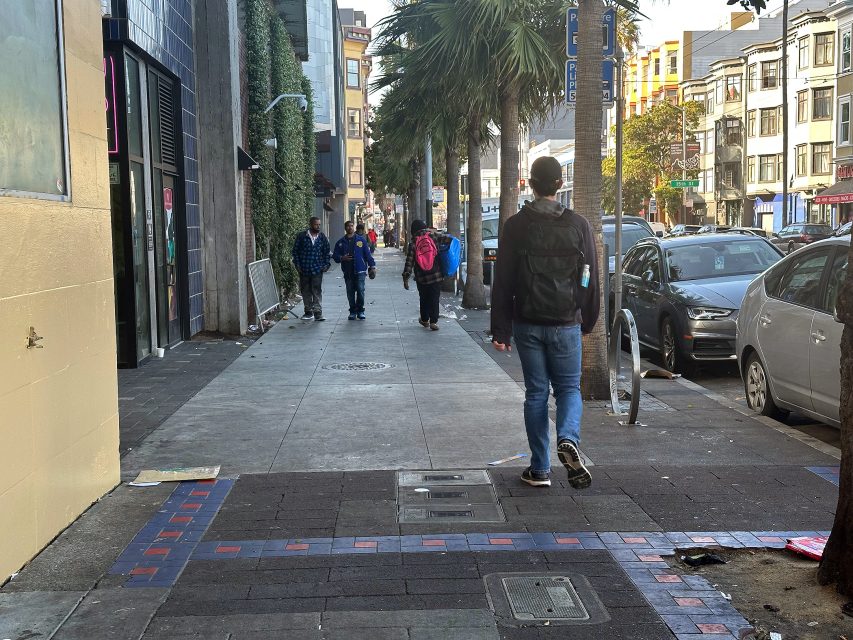 People walk along a city sidewalk lined with parked cars, palm trees, and buildings on a sunny day.