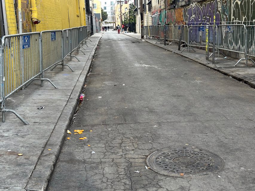 A narrow, cracked alleyway lined with metal barricades and graffiti-covered walls, with a few people visible in the distance.