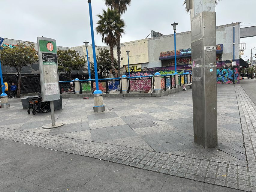 An outdoor urban plaza with gray paving, a few palm trees, blue lamp posts, graffiti-covered walls, and a signpost with a bench and a cart nearby.