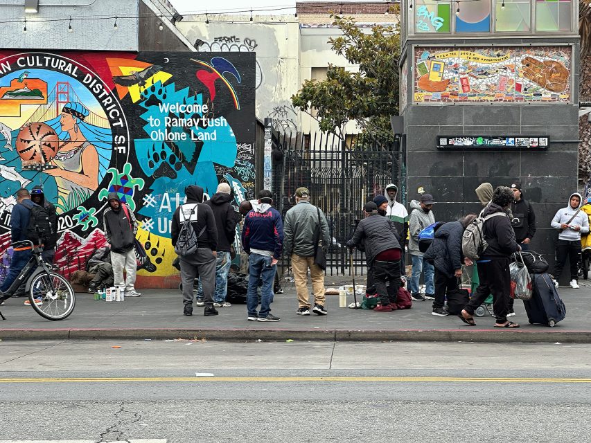 A group of people gather on a city sidewalk in front of a colorful mural and a gated entrance, with various items and backpacks on the ground.