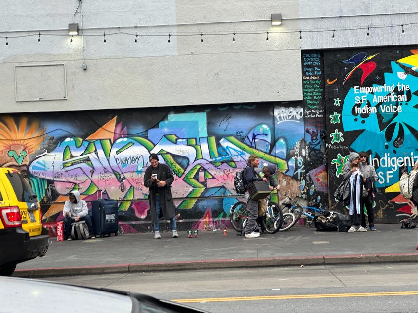 Several people stand or sit with belongings and bicycles on a city sidewalk in front of a colorful mural and a wall with text in San Francisco.