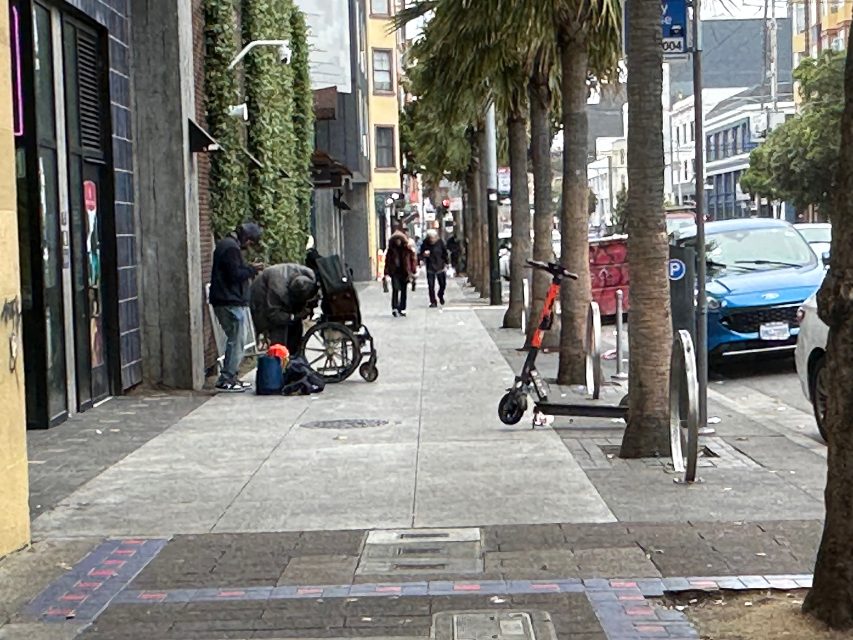 People walk along a city sidewalk; a person with a wheelchair and belongings is on the left. Two electric scooters are parked by palm trees. Cars are parked along the street.