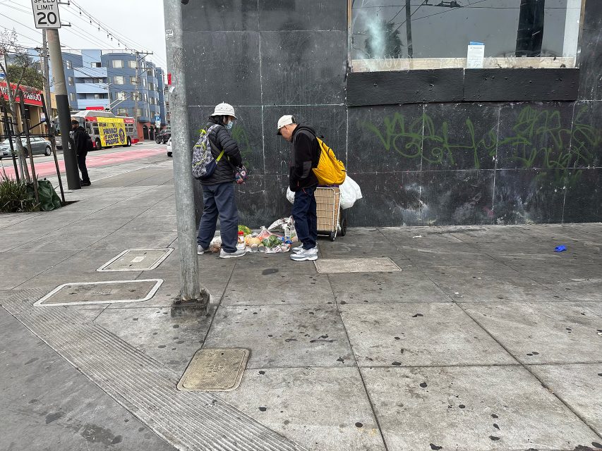 Two people with backpacks and hats stand on a city sidewalk near scattered trash and a dark wall with graffiti.