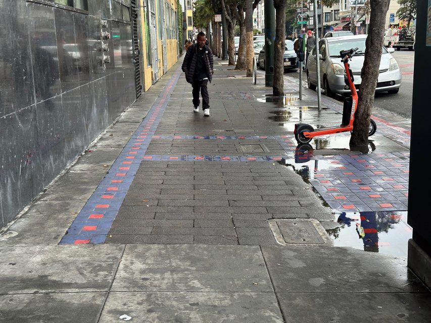 A person walks on a wet city sidewalk with red and blue tile patterns; a parked electric scooter is near a tree, and puddles reflect the surroundings.