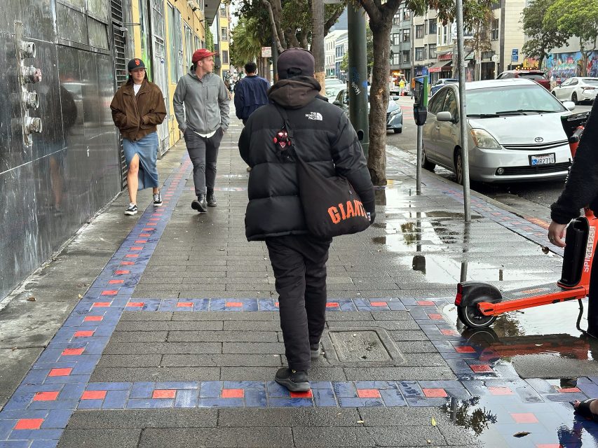 A person in a black puffer jacket carrying a Giants bag walks on a wet city sidewalk; several people walk ahead and a scooter is parked at the curb.