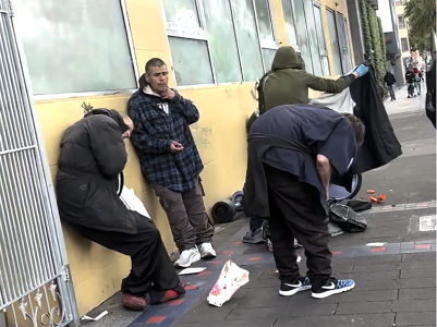 Four people stand and lean against a building on a city sidewalk with scattered belongings and litter around them.