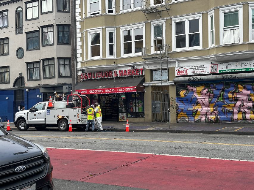 Four workers in safety vests stand near a utility truck on a city street in front of S & N Liquor & Market and a storefront with colorful graffiti. Traffic cones mark their work area.