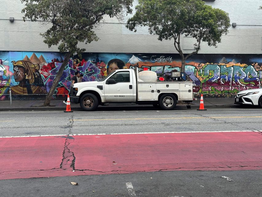 A white utility truck is parked on the street next to orange cones, with colorful graffiti art on the wall behind it and trees on the sidewalk.