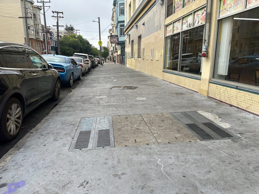 A city sidewalk lined with parked cars on one side and a building with large windows and yellow tiles on the other, with utility covers visible in the pavement.