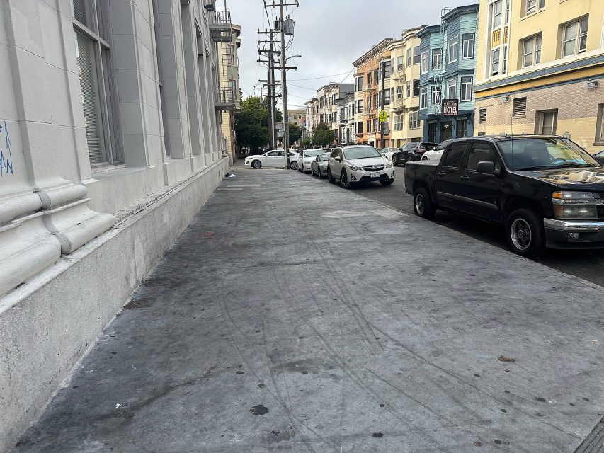 A city sidewalk with tire marks runs alongside parked cars and colorful buildings on an overcast day.