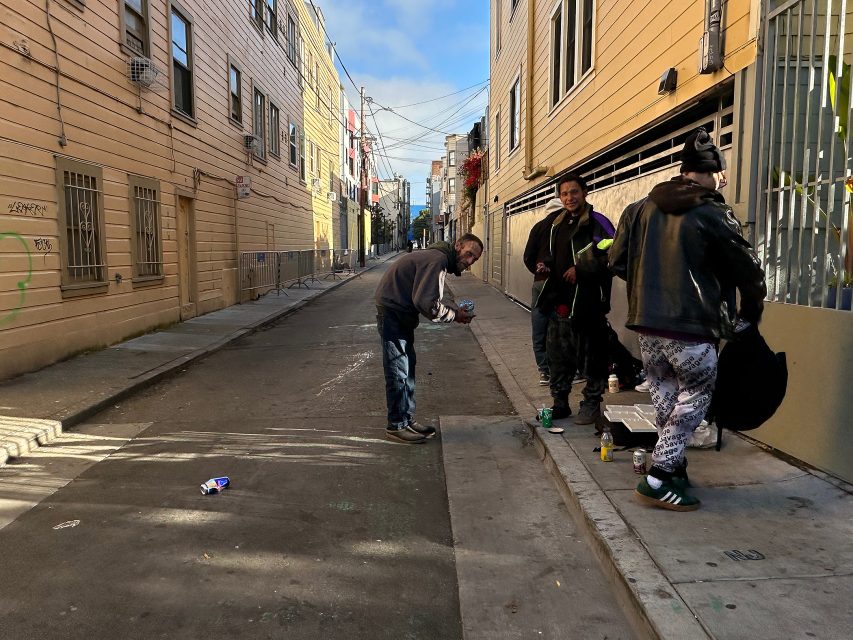 A man bends down in a narrow urban alley as three others stand nearby on the sidewalk; various items are scattered on the ground.