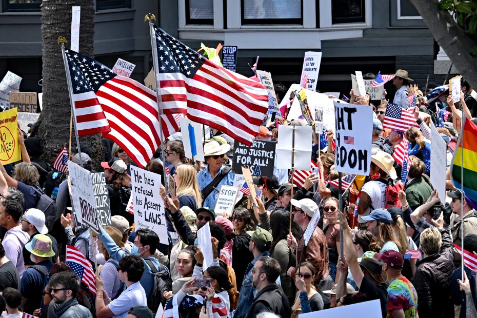 A large crowd of people hold American flags and protest signs while gathered outdoors during a daytime demonstration.