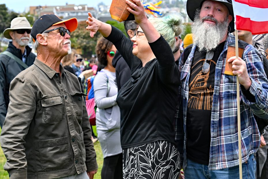 A group of people stand outdoors at a gathering; one woman raises her arms holding an object while others watch, and a man holds a red and white flag.