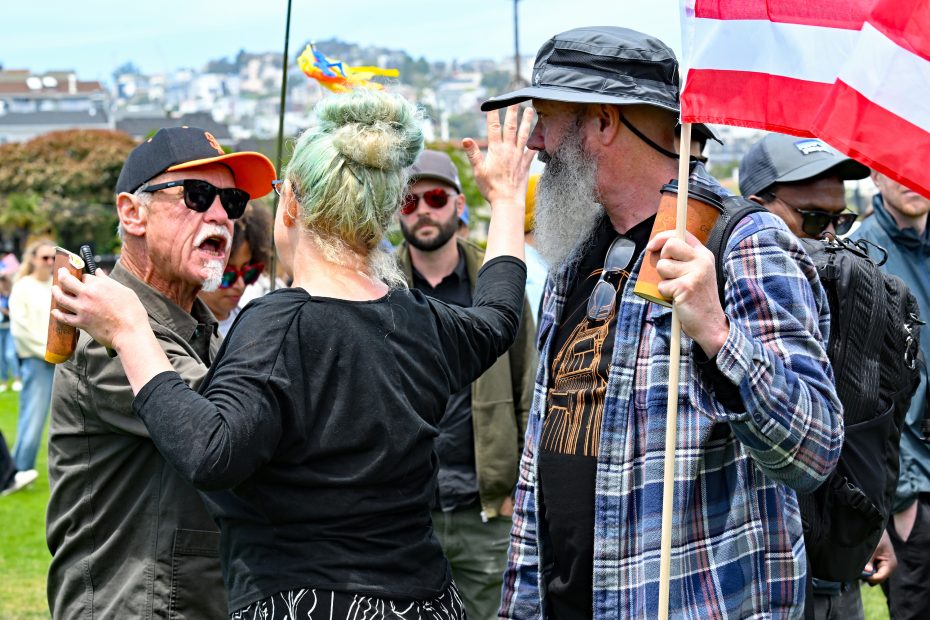 Three people argue outdoors; one woman raises her hand between two men, one holding a drink and the other holding an American flag. Several people watch in the background.
