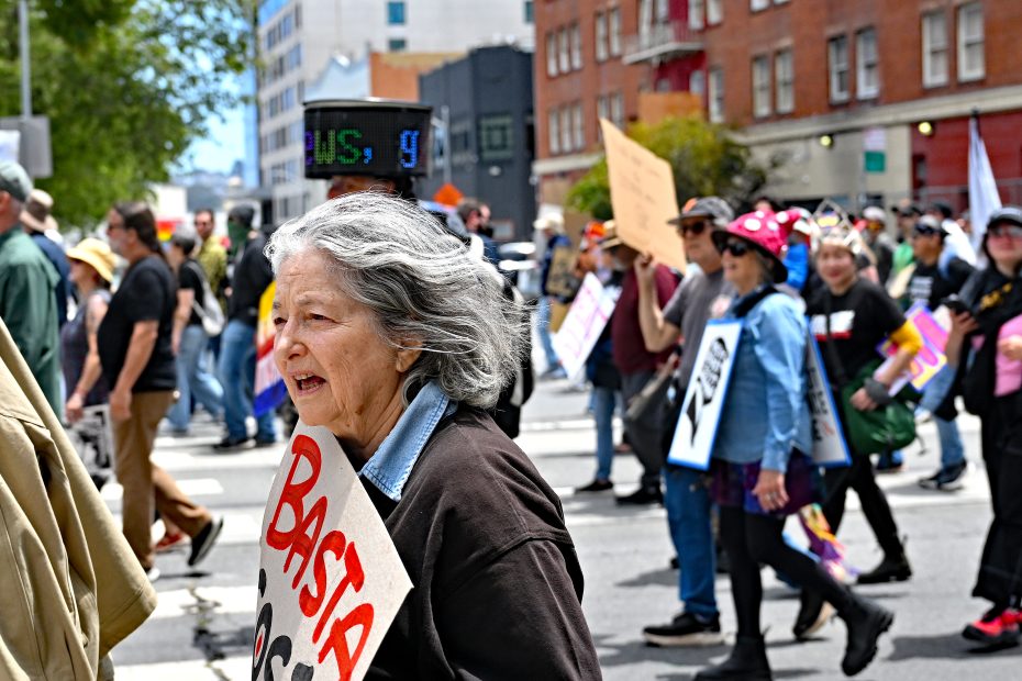 An older woman holds a protest sign that reads "BASTA" while marching with a crowd of demonstrators on a city street.