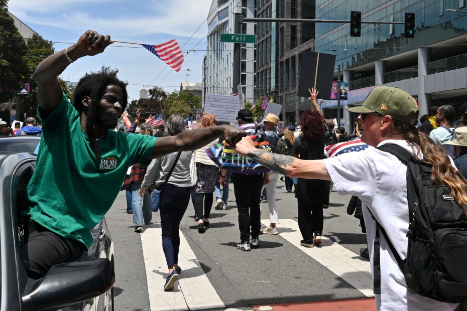 A man in a car hands a small American flag to a woman as a crowd marches along a city street holding signs.