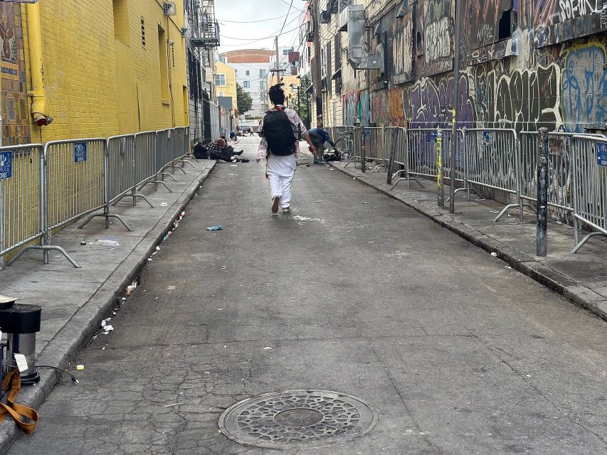 A person walks down a graffiti-covered alley lined with metal barricades and scattered trash; several people sit by the walls in the background.