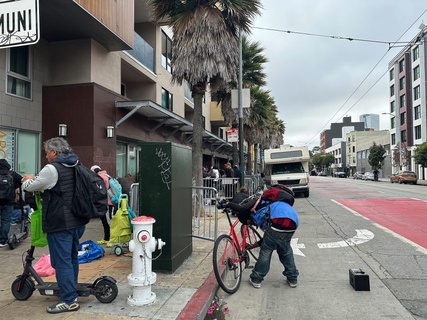 A person repairs a bicycle on a city sidewalk while others stand nearby; a row of palm trees, buildings, and a parked RV are visible in the background.