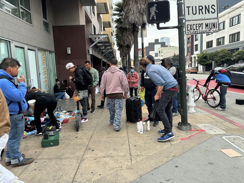 A group of people stand and browse items laid out on the sidewalk near a street corner in an urban area.