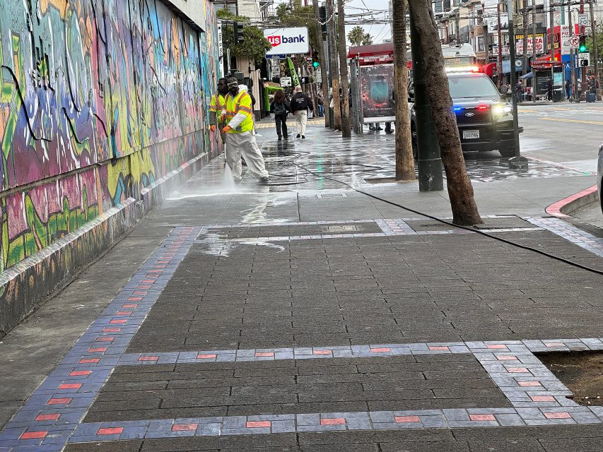 A worker in a reflective vest uses a power washer to clean a graffiti-covered wall on a city sidewalk; a police car with flashing lights is parked nearby.