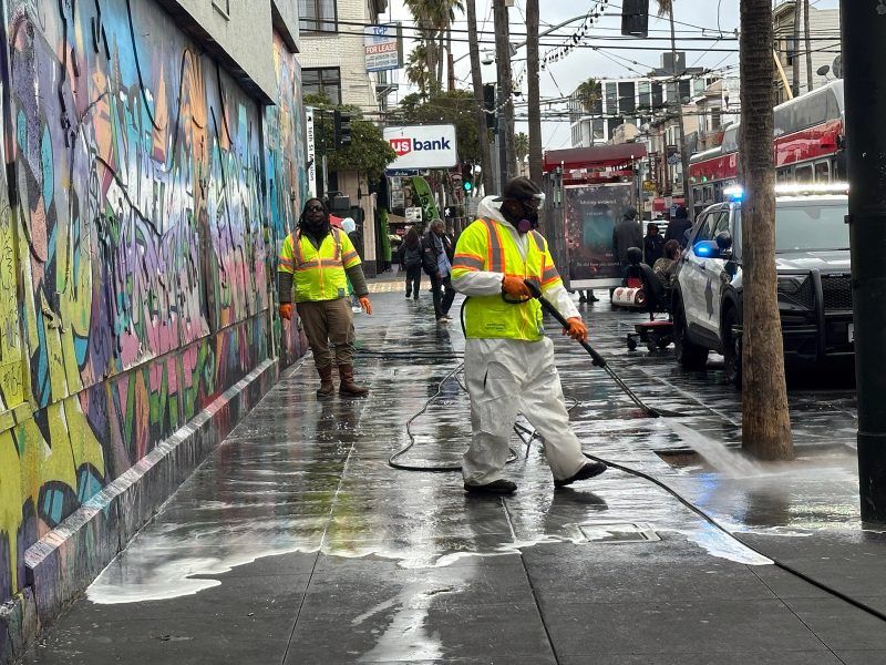 Workers in protective gear power-wash a graffiti-covered city sidewalk; police cars with lights on are parked nearby on a wet street.