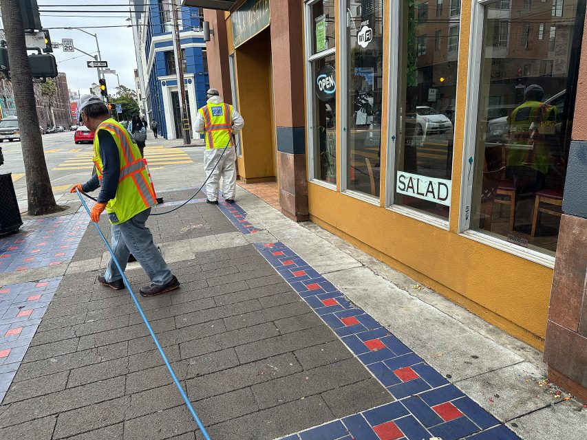 Two workers in safety vests and gloves clean a city sidewalk outside a yellow building with a "SALAD" sign in the window.