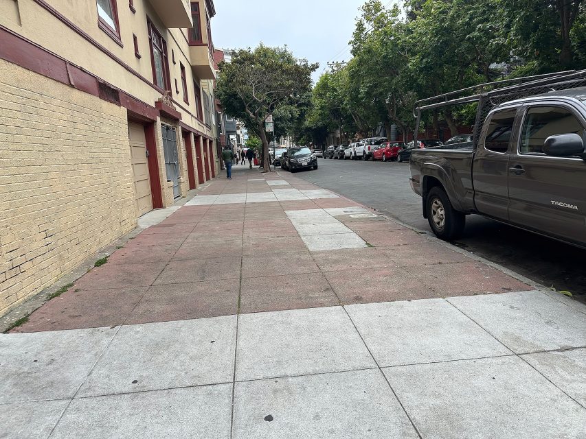 A mostly empty city sidewalk runs beside parked cars and apartment buildings, with a few pedestrians visible in the distance under overcast skies.