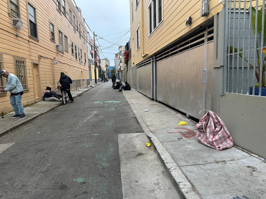 A narrow urban alley with several people sitting or standing along the sides, some belongings and litter scattered on the ground.
