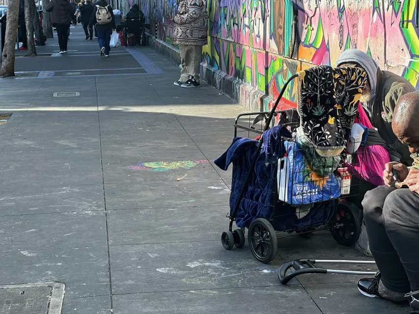 A person sits on a city sidewalk beside a walker and a cart filled with belongings, near a colorful graffiti-covered wall. Pedestrians walk in the background.