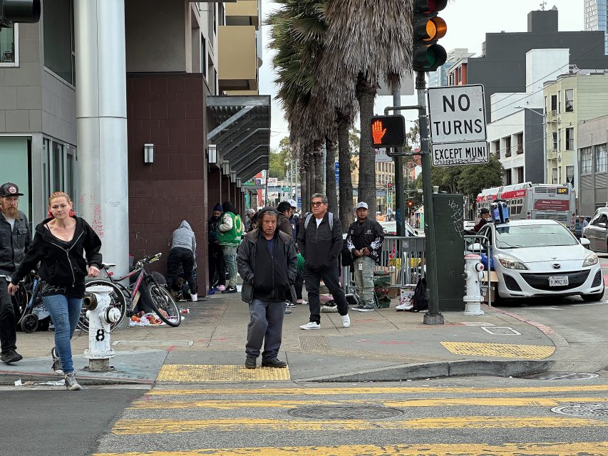 A busy urban street corner with people standing and walking, bicycles, a “No Turns” sign, and traffic lights; buildings and cars are visible in the background.
